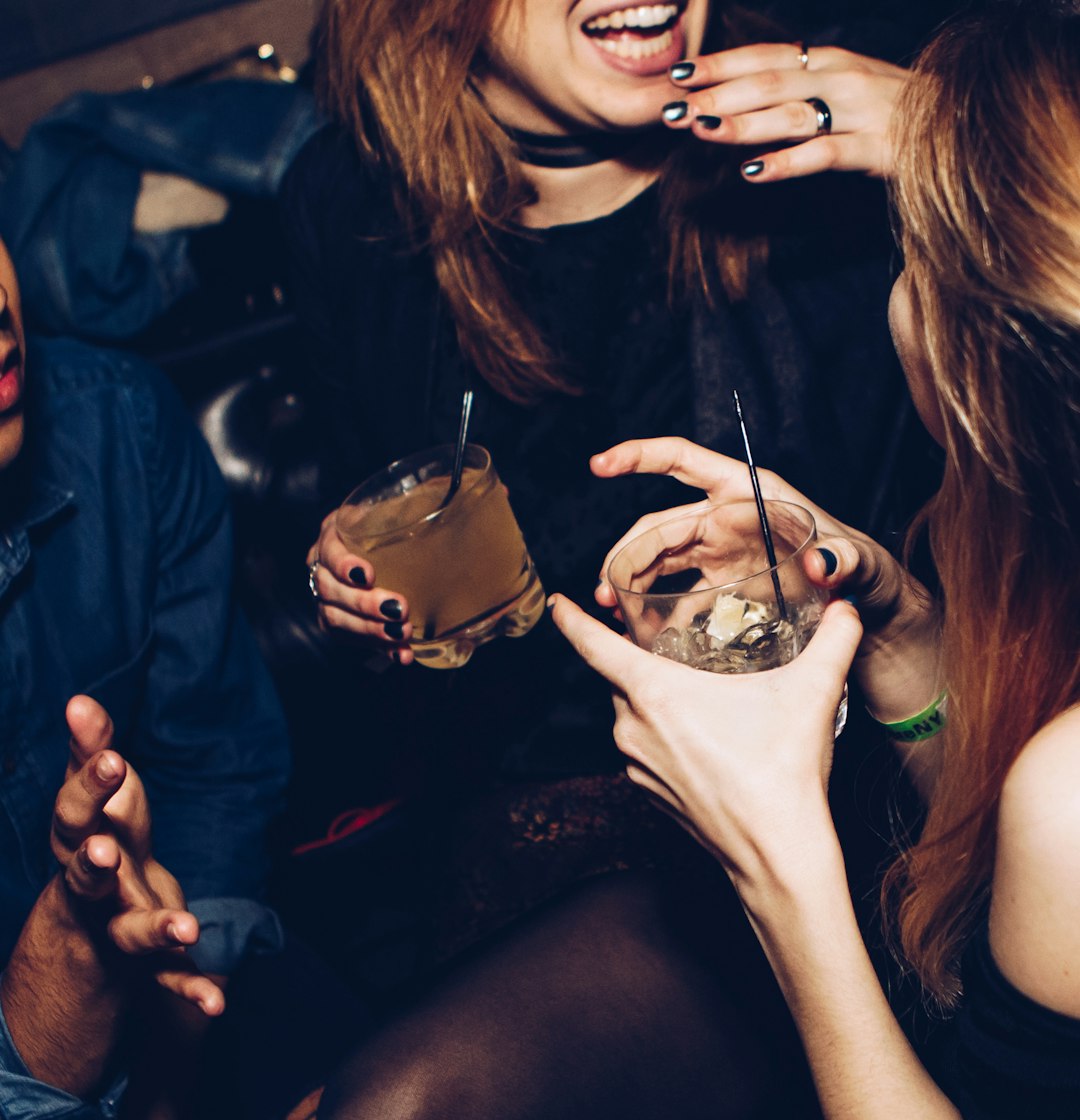 Group of friends enjoying drinks and laughing together in a dimly lit social setting, holding cocktails with ice and straws.