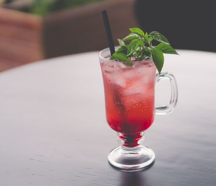 A red, leafy adaptogenic drink with ice and a straw in it, sitting on a tabletop with blurred background.