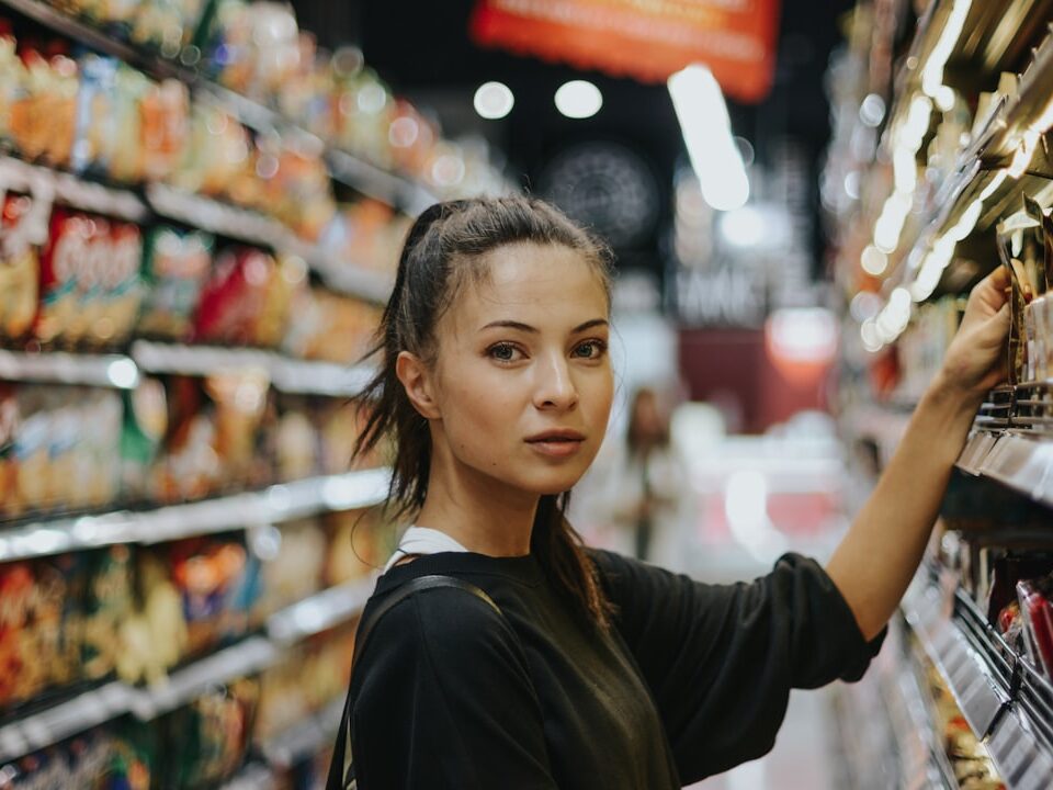 Young woman shopping in a brightly lit grocery store aisle, reaching for a product on the shelf while surrounded by colorful snack displays.