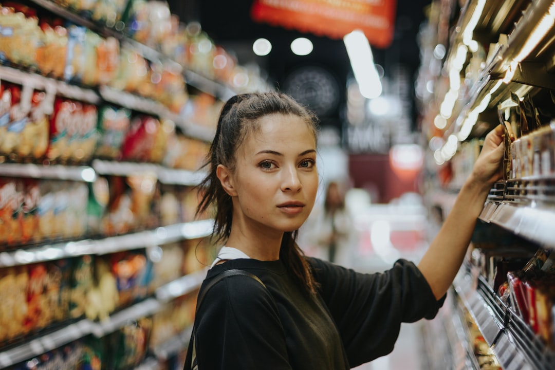 Young woman shopping in a brightly lit grocery store aisle, reaching for a product on the shelf while surrounded by colorful snack displays.